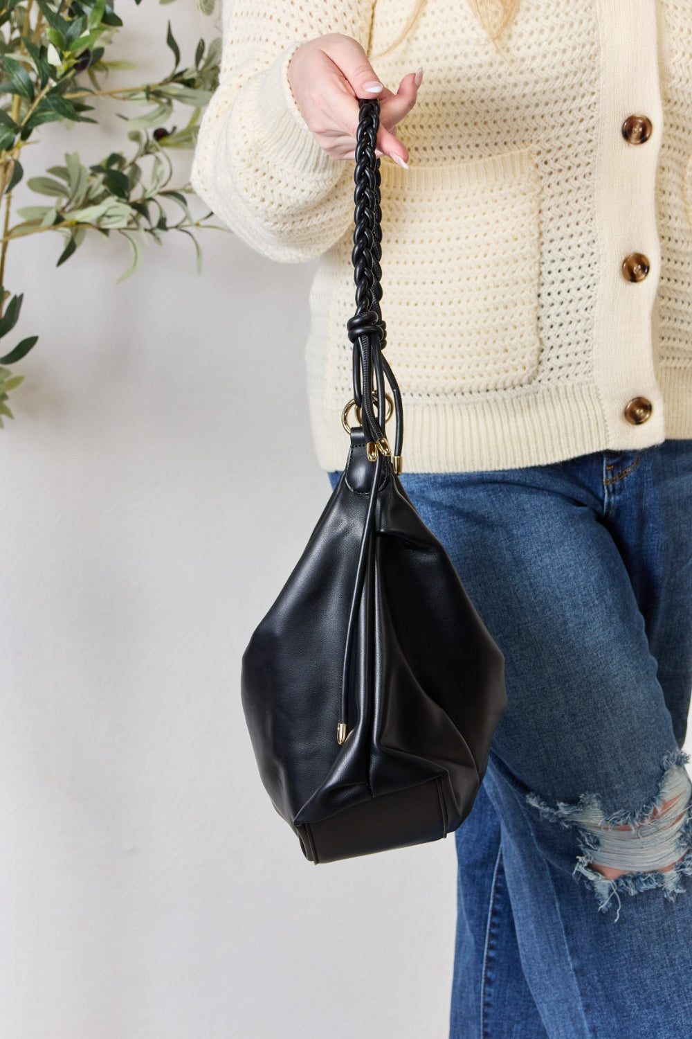 A person in a knitted sweater and jeans holds the SHOMICO Braided Strap Shoulder Bag, a blue-green statement accessory featuring straps crafted from vegan leather.