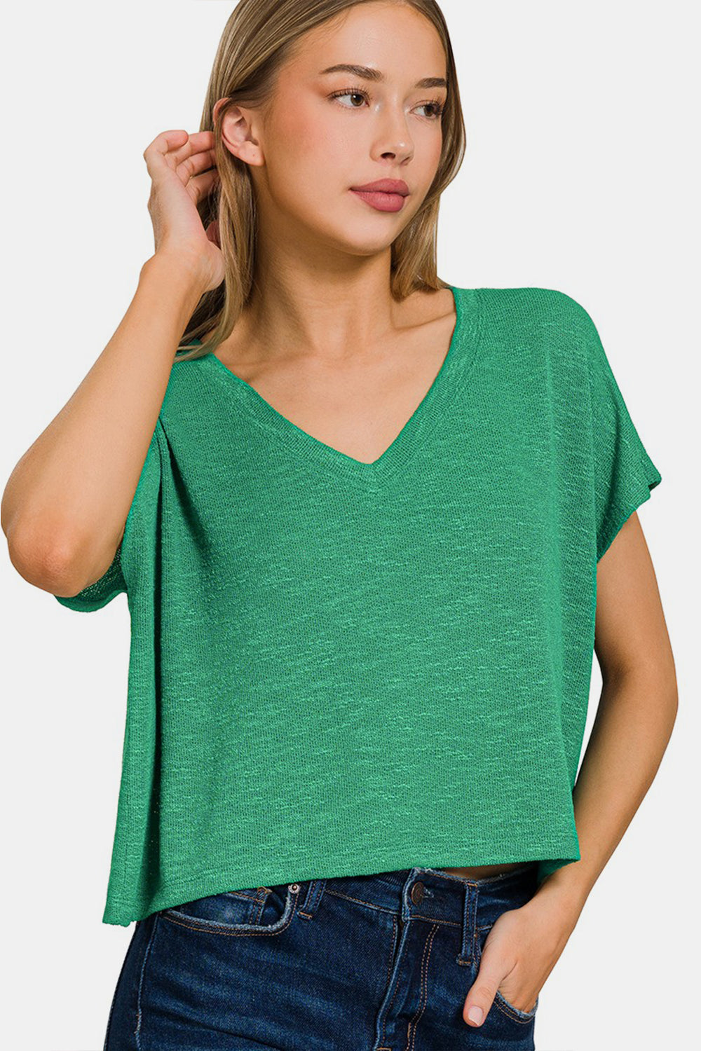 A person with long hair wears a green Zenana V-Neck Short Sleeve T-Shirt and blue jeans, standing against a white background. This versatile wardrobe essential offers both a stylish look and comfortable fit.