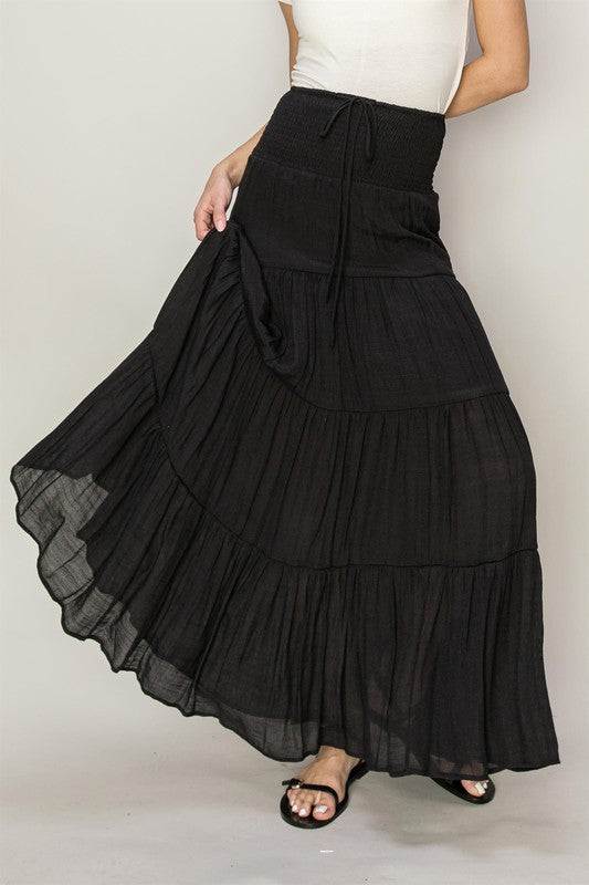 A person wearing the D-Drawstring Waist Tiered Maxi Skirt in black, featuring a subtle boho print, paired with a white top, with one leg stepping forward.