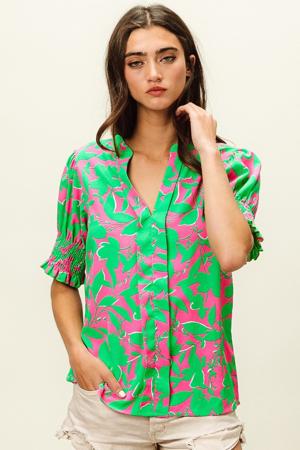 A woman wearing the BiBi Smocked Cuff Floral Contrast Top, featuring a chic design with bright pink and green floral patterns and ruffled sleeves, poses with one hand near her chin.