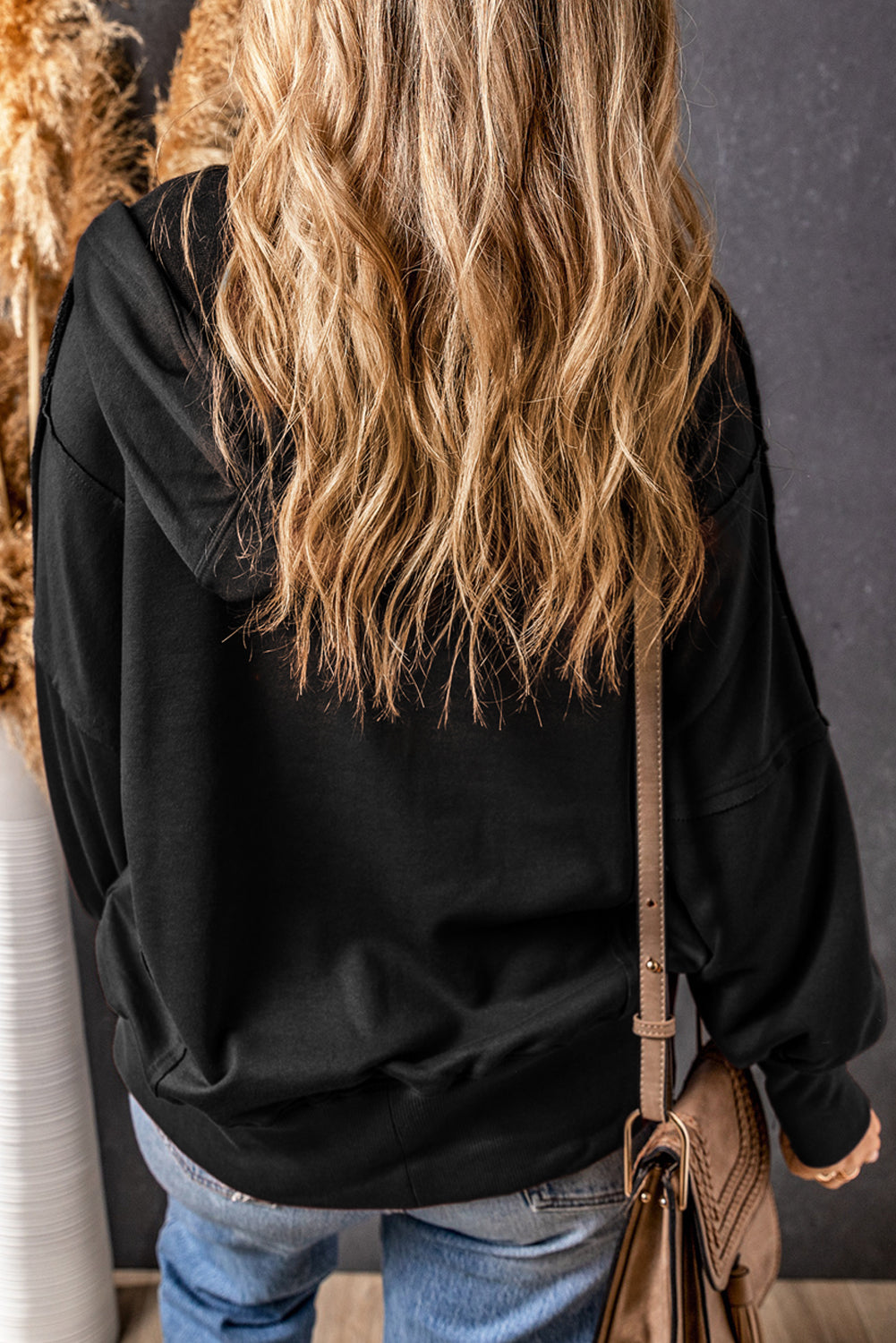 A person with long, wavy blonde hair is shown from the back, wearing a Black Raw Edge Exposed Seam Full Zip Hoodie and blue jeans, and carrying a beige shoulder bag.