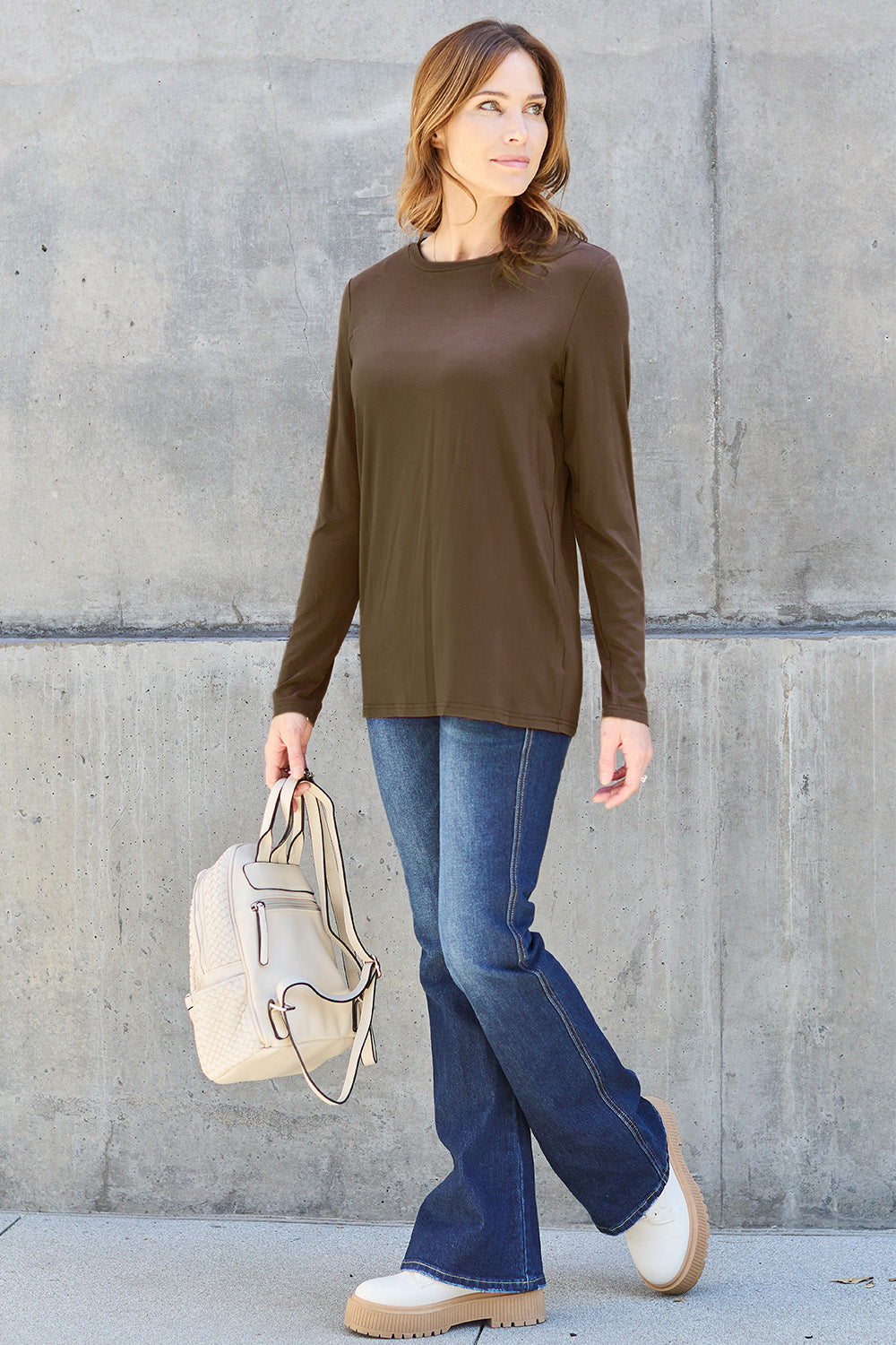 A woman with shoulder-length hair wearing the Basic Bae Full Size Round Neck Long Sleeve Top and blue jeans stands in front of a concrete wall, holding a beige handbag over her shoulder. Her outfit exemplifies basic style, with the top made from 100% polyester and recommended for machine wash cold.