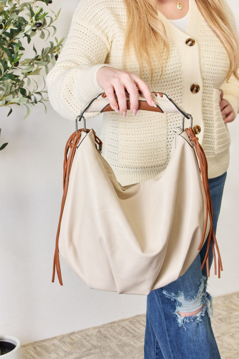 Individual holding a SHOMICO Fringe Detail Contrast Handbag, featuring cream and brown accents, paired with a knitted cream sweater, standing against a light background. The handbag exudes Bohemian flair with its relaxed style.