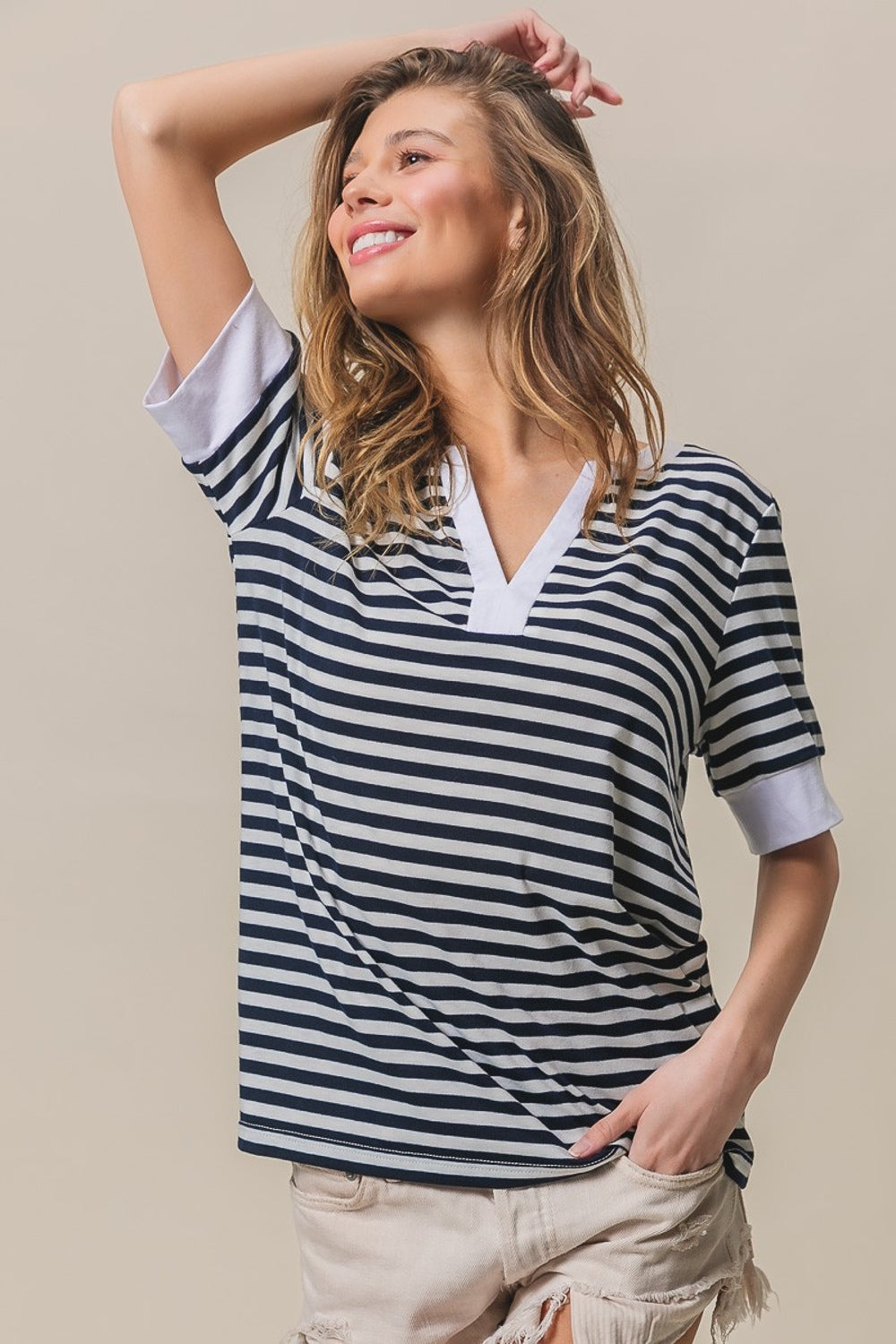 A person with long hair, wearing a BiBi Contrast Striped Notched Knit Top in navy and white and beige shorts, is posing with one hand in their hair, smiling against a beige background.