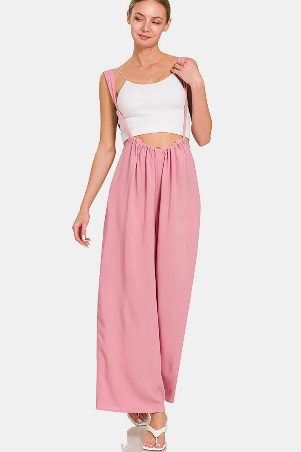 A woman is smiling in her fashion-forward style, donning the Zenana Pocketed Wide Strap Wide Leg Overalls in pink along with a white crop top. She has her hands in the overall's pockets and complements the look with white high-heeled shoes.