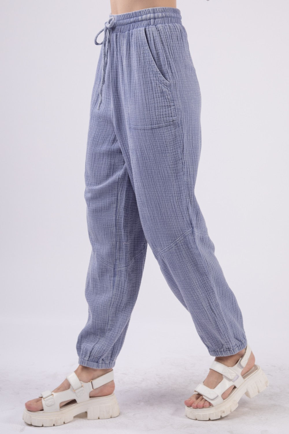 A person wearing VERY J Washed Woven Crinkle Gauze Drawstring Cargo Pants in light blue, paired with white sandals. These relaxed-fit, textured pants feature an elastic waistband and pockets, showcasing a utility design that's perfect for blending comfort and functionality against a plain background.