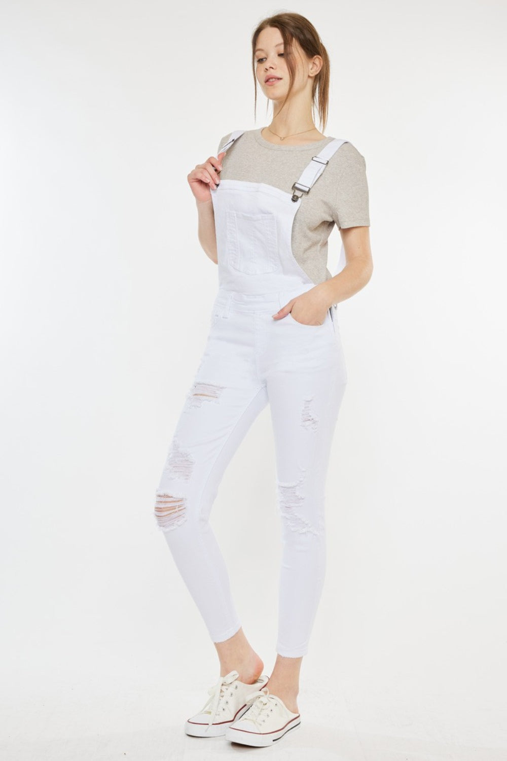 A fashion-forward woman stands against a plain background, wearing a grey t-shirt, Kancan Distressed Skinny Denim Overalls in white, and white sneakers with red detailing.