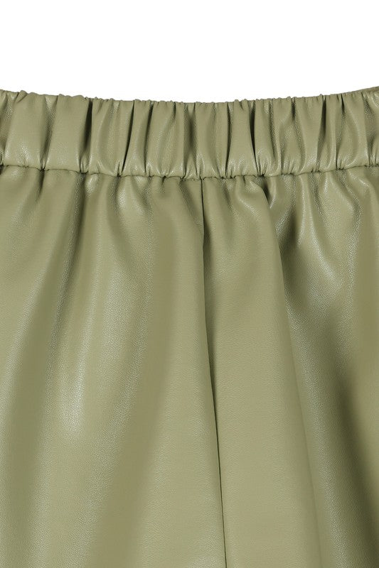 An individual is dressed in a light green sweater paired with pleated vegan leather shorts, featuring a functional pocket where one hand rests.