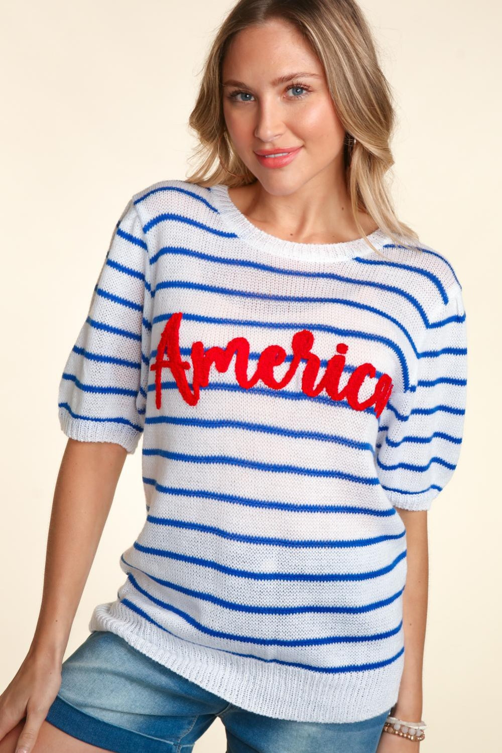 A woman wearing a chic Haptics Letter Embroidery Striped Knit Top featuring "America" in red stands against a neutral background. The white and blue striped knit design pairs perfectly with her denim shorts.