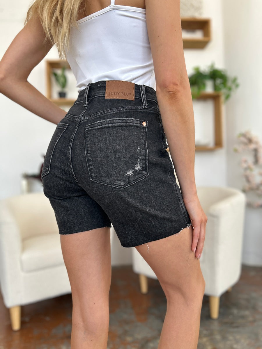 A person wearing a white t-shirt and Judy Blue Full Size High Waist Rigid Magic Denim Shorts is standing indoors in front of a beige chair.