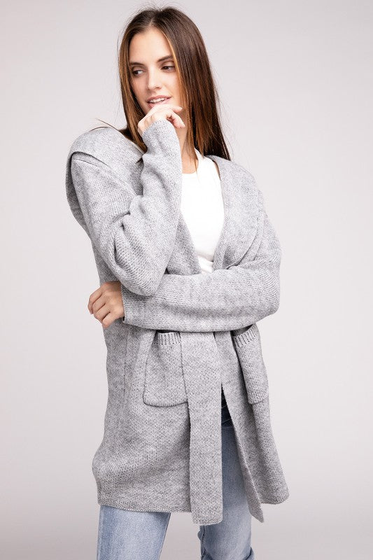 A woman modeling a cozy Hooded Open Front Sweater Cardigan over a black top, posing with one hand in her pocket against a gray background.