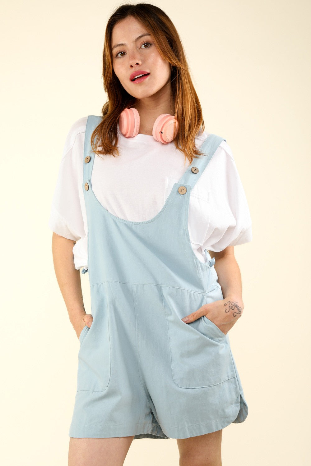 A person stands against a light background, wearing a white shirt, VERY J Adjustable Waist Suspender Overalls with Pockets, and pink headphones around their neck.