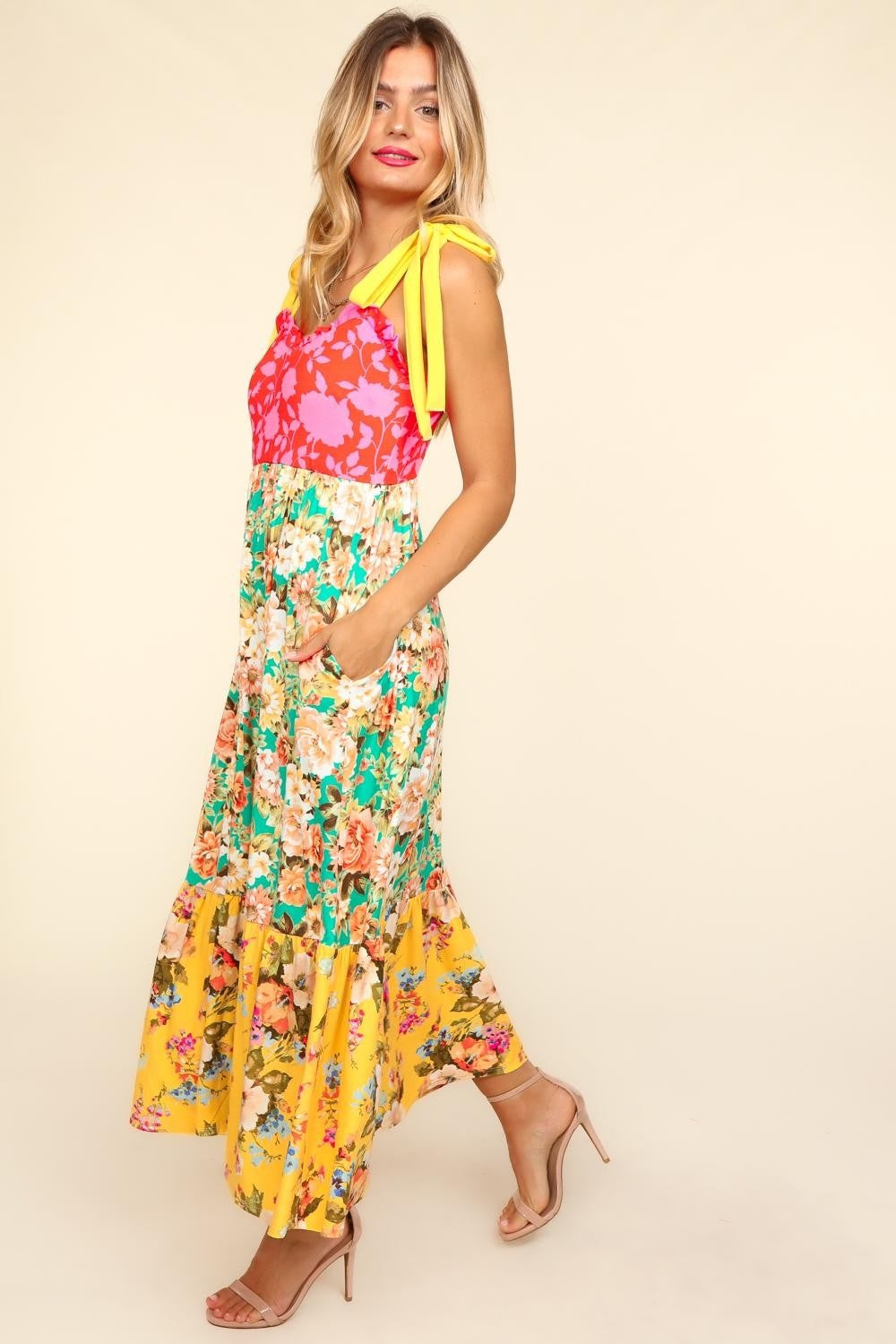 A person wearing the Haptics Floral Color Block Maxi Dress with Pockets, featuring a vibrant floral print and a color block maxi skirt in pink and yellow hues, stands smiling against a beige background.