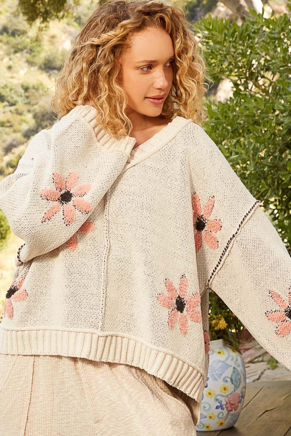 A person with curly hair smiles while wearing a cozy POL V-Neck Floral Pattern Chenille Sweater. They stand outdoors on a wooden deck with greenery and a ceramic vase in the background.