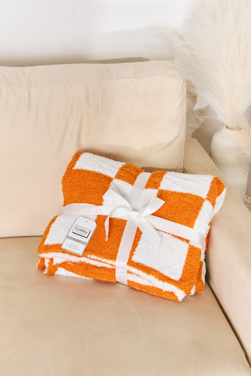 A person stands holding a large Cuddley Checkered Decorative Throw Blanket, smiling, in a room with a sofa and a potted plant. The blanket features a black and white checkerboard pattern, is made from 100% polyester, and imported for quality.