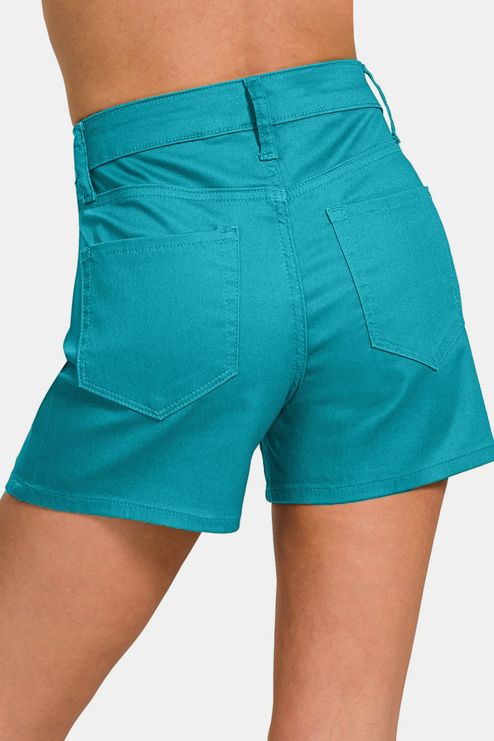 A person is wearing Zenana High Waist Denim Shorts in teal with hands partially in the pockets. The shorts, featuring a button and zipper closure, offer a flattering fit and are perfect for any summer wardrobe.