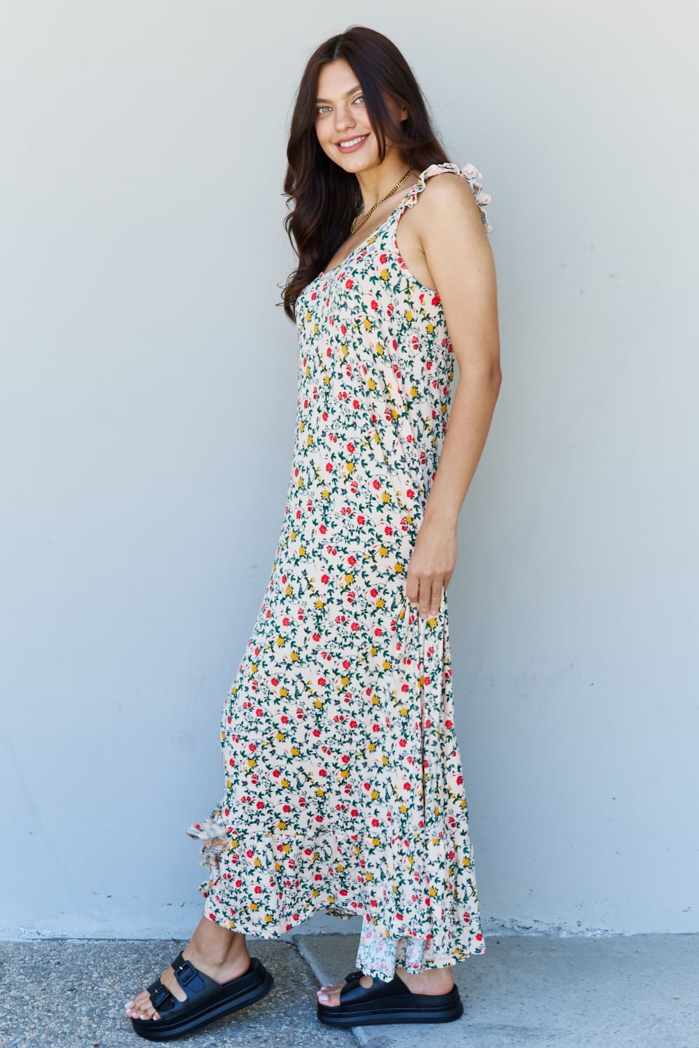 A woman is standing against a gray wall, wearing the Doublju In The Garden Ruffle Floral Maxi Dress in Natural Rose and black sandals, with her left hand raised to her head.