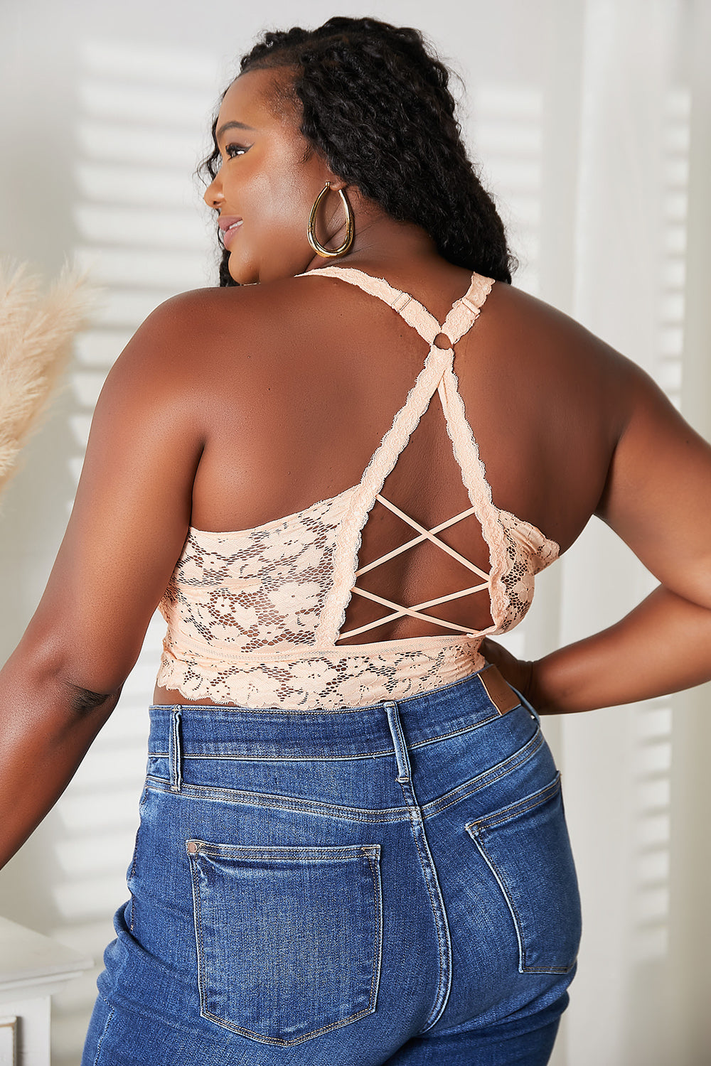 A woman with long blonde hair, wearing a JadyK Juliette Full Size Crisscross Lace Bralette in peach and blue jeans, stands in front of a neutral background with dried plants.