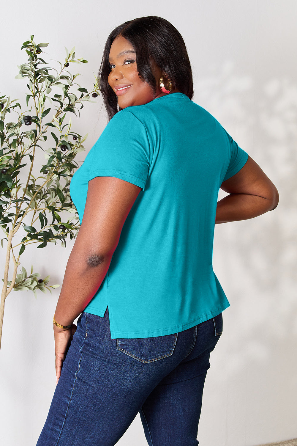 A woman with long hair, smiling with her right hand in her pocket and standing next to a plant, is wearing the Basic Bae Full Size Round Neck Short Sleeve T-Shirt in black along with beige pants made of rayon spandex.