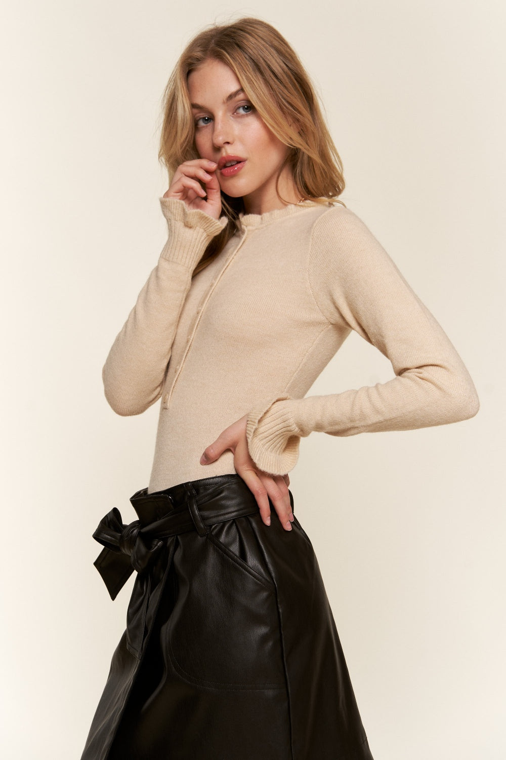 A woman in a beige sweater and black leather skirt stands against a neutral background, her look perfectly complemented by the sleek lines of the And The Why Half Button Round Neck Long Sleeve Bodysuit underneath.