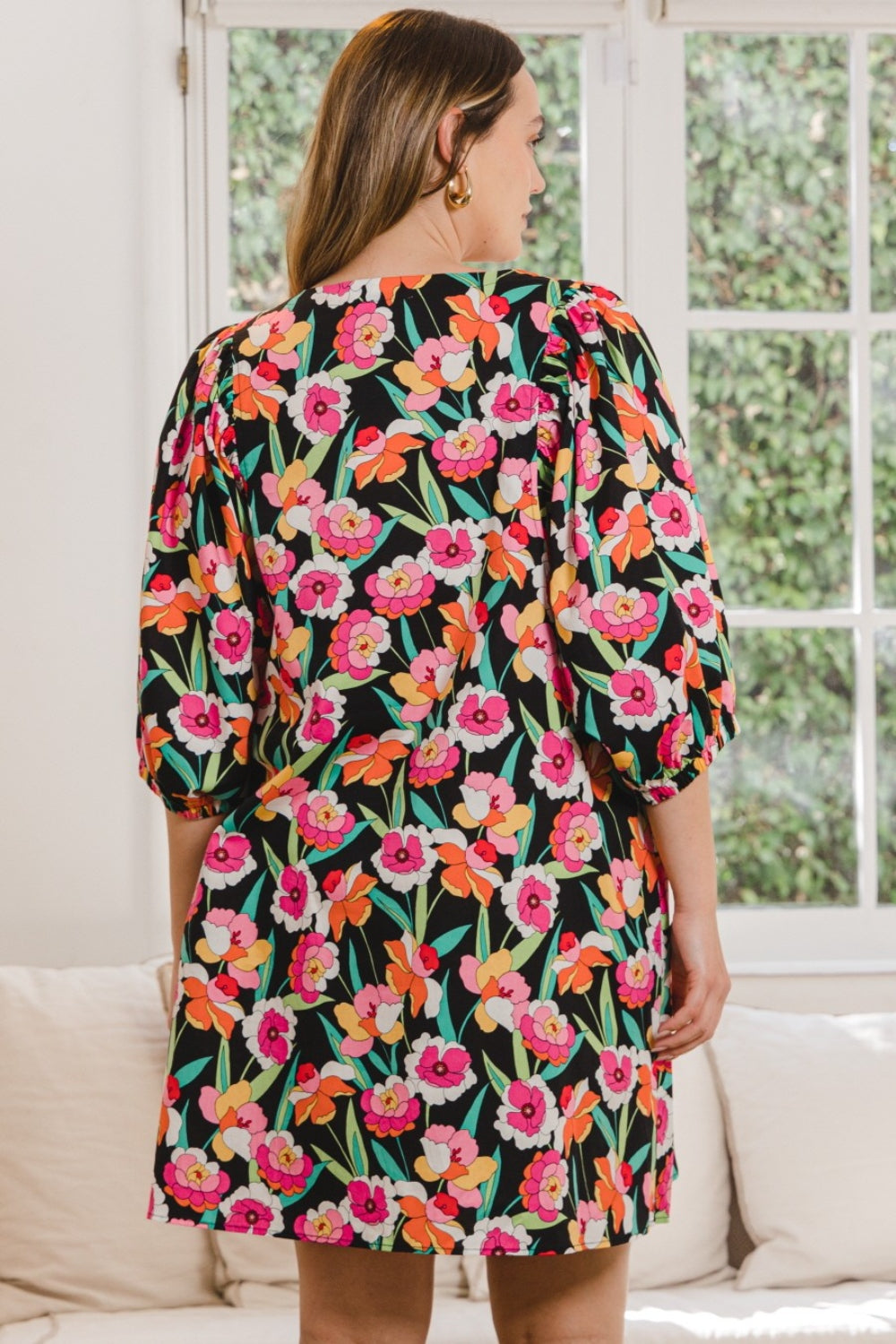 A woman wearing the stylish ODDI Full Size Floral Puff Sleeve Mini Dress stands indoors, framed by a large window with lush greenery outside.