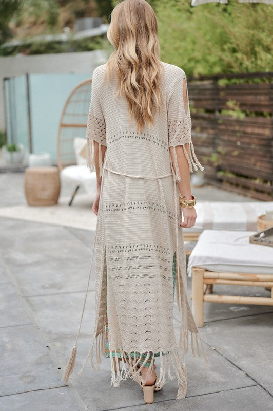 A woman in a green floral dress and a Solid Long Cardigan With Fringe exudes boho-chic vibes as she strolls on a patio, surrounded by outdoor furniture and lush plants.