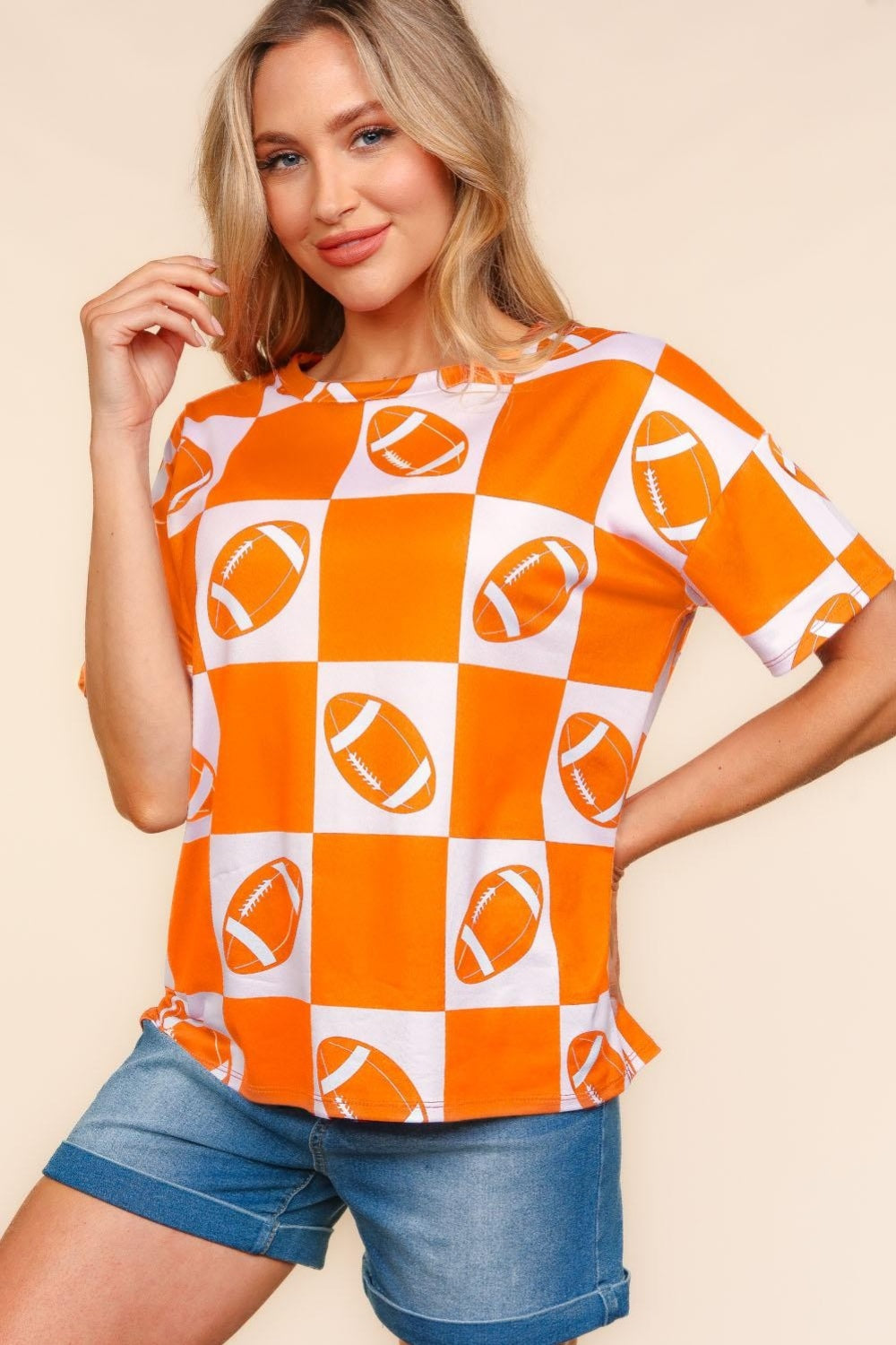 A woman wears a Haptics Football Checkered Print Short Sleeve T-Shirt in orange and white, standing against a plain background, and pairs it perfectly with denim shorts for a casual sporty look.