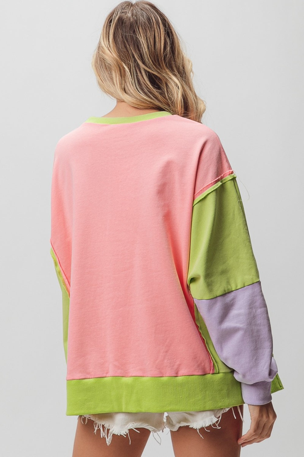 A woman is wearing a BiBi Washed Color Block Sweatshirt featuring vintage-inspired sections in pink, green, and purple. She has paired it with white distressed shorts and is posing with one arm slightly raised, perfectly showcasing the relaxed fit.