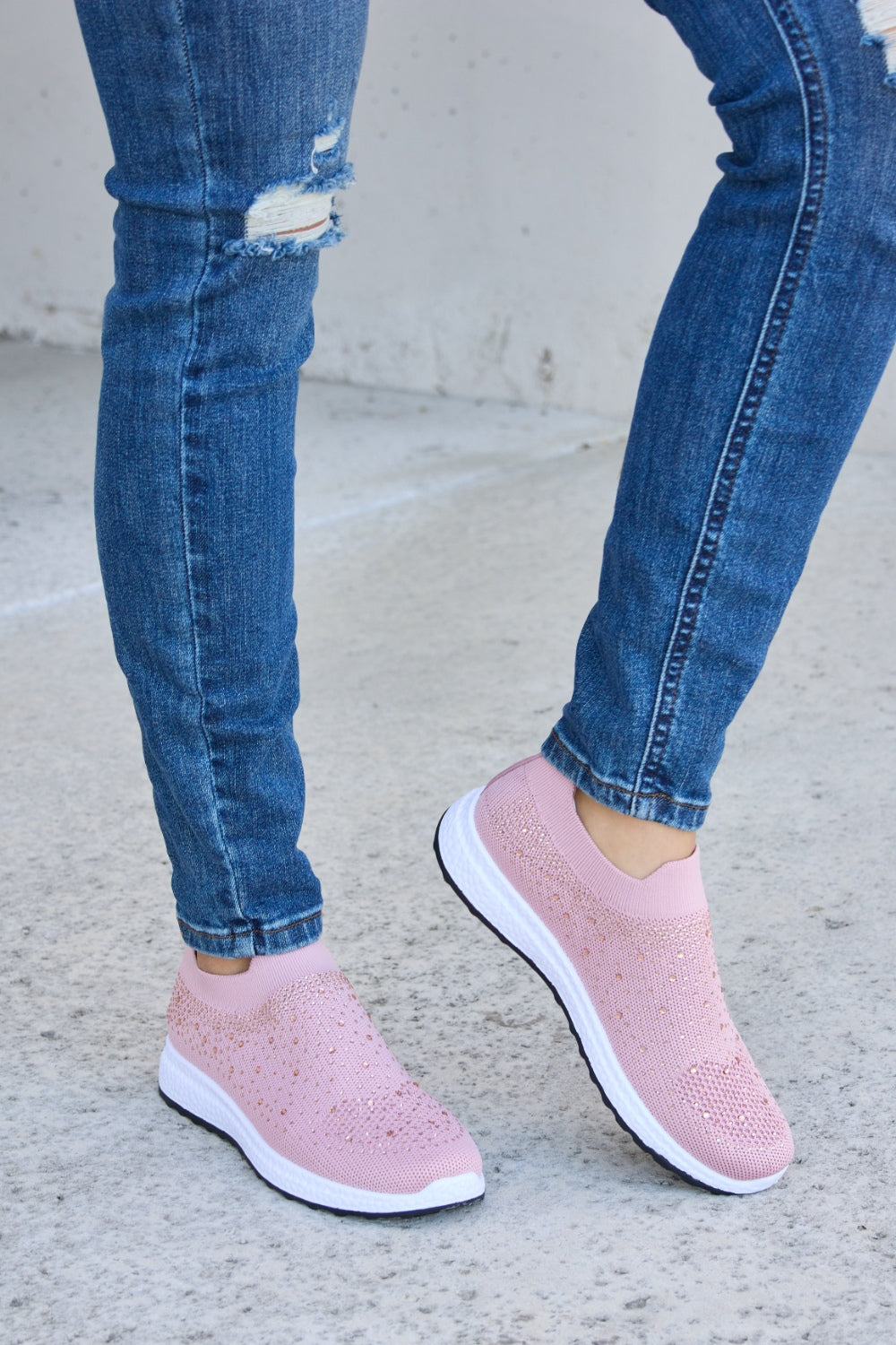 Person wearing Forever Link Rhinestone Mesh Breathable Sneakers in light pink and ripped blue jeans, standing on a concrete surface.