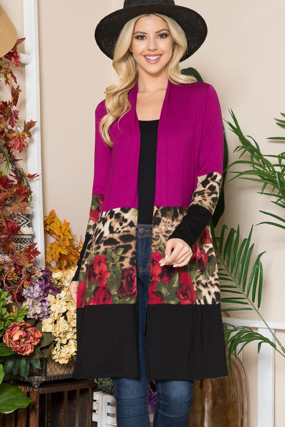 A person wearing a hat and the Celeste Full Size Color Block Open Front Cardigan, featuring magenta, leopard print, and floral designs, stands indoors amidst an array of colorful flowers and plants.