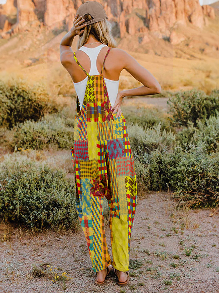 A woman stands outdoors in front of a rocky landscape, wearing the Full Size Printed V-Neck Sleeveless Jumpsuit and a brown brimmed hat. She is smiling and holding the brim of her hat with one hand, exuding the carefree vibe of this vibrant and colorful jumpsuit.