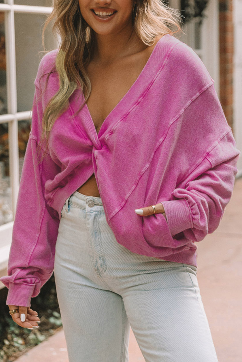 A woman stands with her back to the camera, wearing a long-sleeved Rose Exposed Seam Twist Open Back Oversized Sweatshirt and light blue jeans, showcasing a relaxed fit.