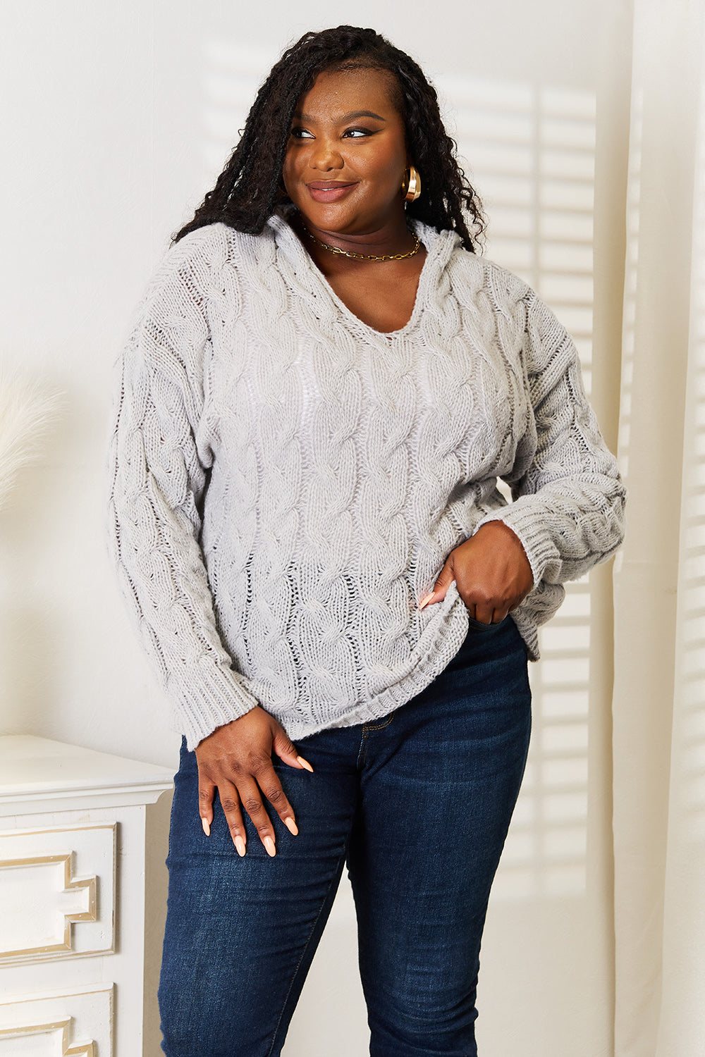 A person with long hair wearing a cozy Woven Right Cable-Knit Hooded Sweater and dark jeans is standing indoors looking to the side.