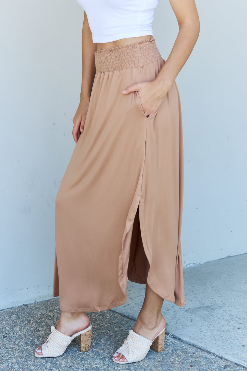 A person stands against a wall, wearing a white sleeveless top, the Doublju Comfort Princess Full Size High Waist Scoop Hem Maxi Skirt in Tan, and blue platform sandals. The right hand rests on the side of the skirt.