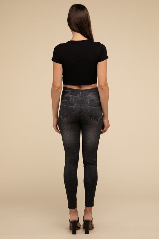 A person wearing Denim Leggings in a high-waisted black design and paired with black heels poses against a plain beige background, highlighting the sleek, stretchy fit.