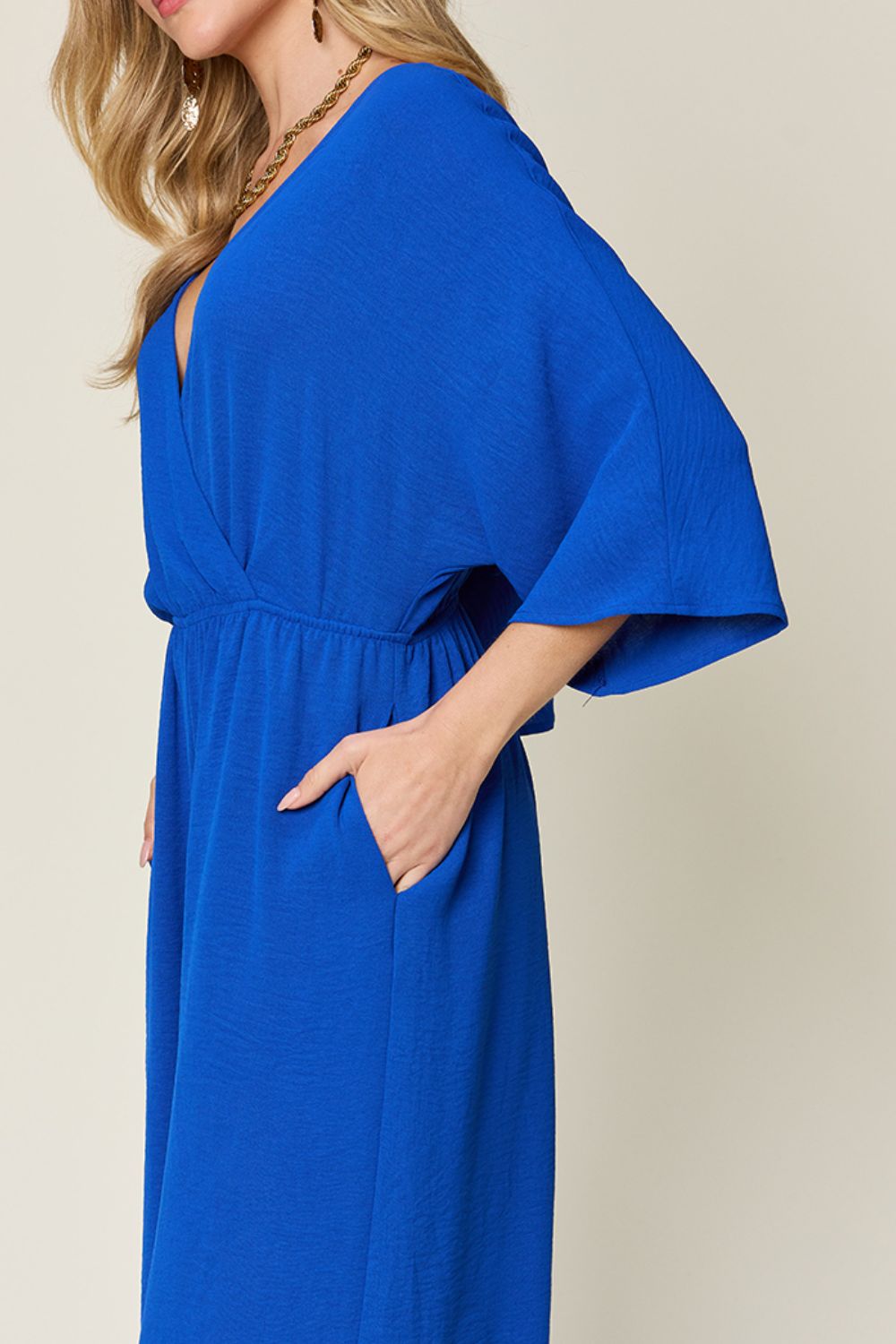 Person wearing the Double Take Full Size Surplice Wide Leg Jumpsuit with Pockets in royal blue, featuring wide legs, a deep V-neckline, and ruched detailing, posing with hands on hips against a plain light-colored background.