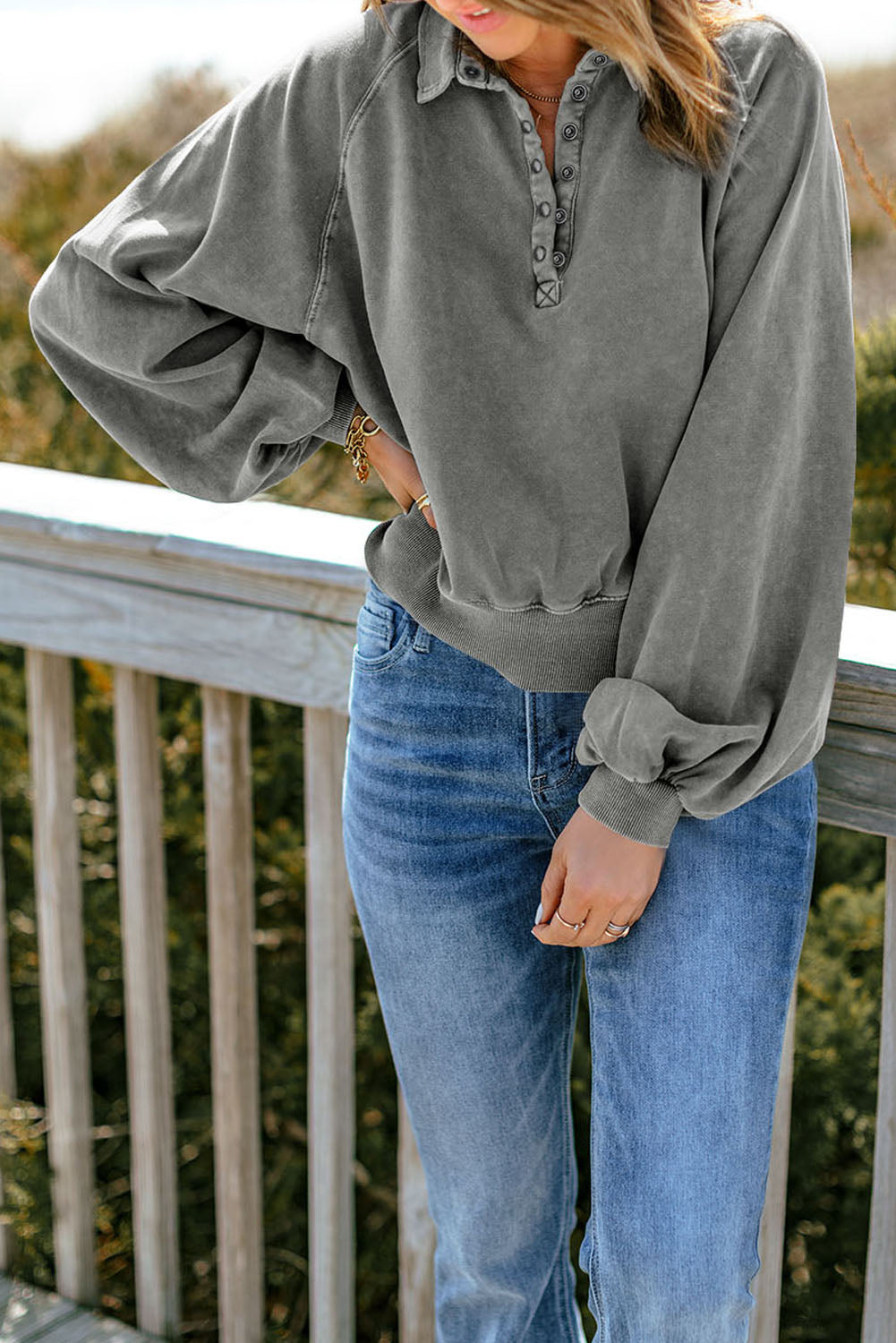 A person with long hair stands outdoors, facing away, wearing a chic **Gray Washed Snap Buttons Lantern Sleeve Pullover Sweatshirt** and blue jeans, resting their hands on a wooden railing.