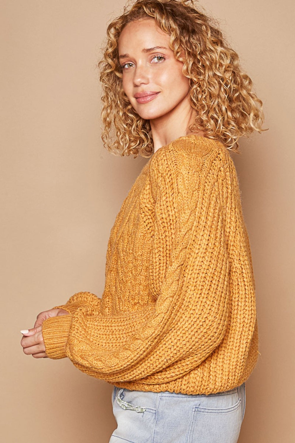 A person with curly hair wears a POL Cable Knit Cutout Long Sleeve Sweater in mustard, paired with light blue jeans adorned with patchwork designs.