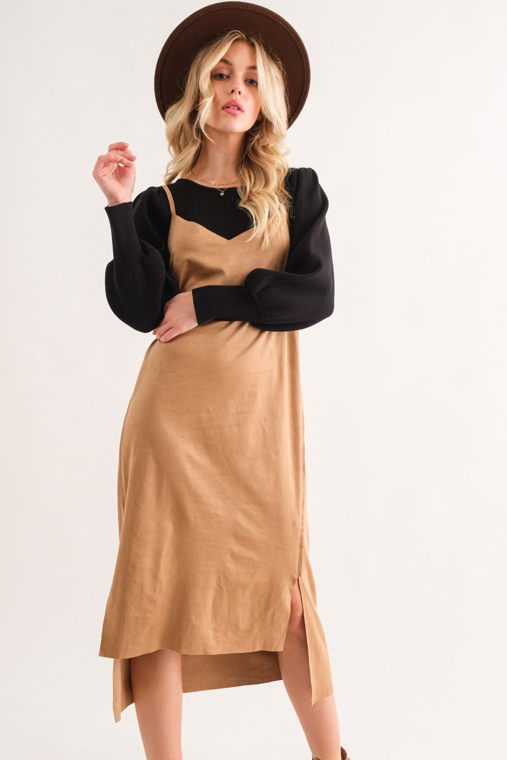 A woman wearing a wide-brimmed hat, black long-sleeve top, and the And The Why Faux Suede Cut Edge Slit Cami Midi Dress poses against a plain background.