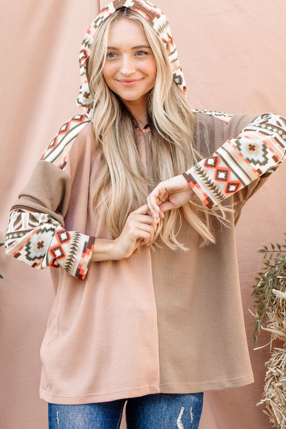 A woman with long blonde hair wears an And The Why Printed Color Block Hooded Waffle Top in various earth tones, standing in front of a light brown background.