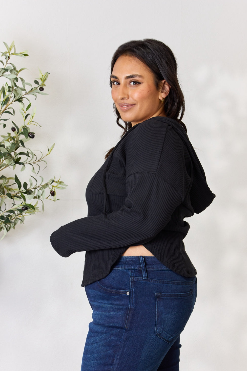 A person wearing the Culture Code Full Size Ribbed Zip Up Drawstring Hooded Jacket and blue jeans stands in front of a plain background with a small leafy plant to the left.