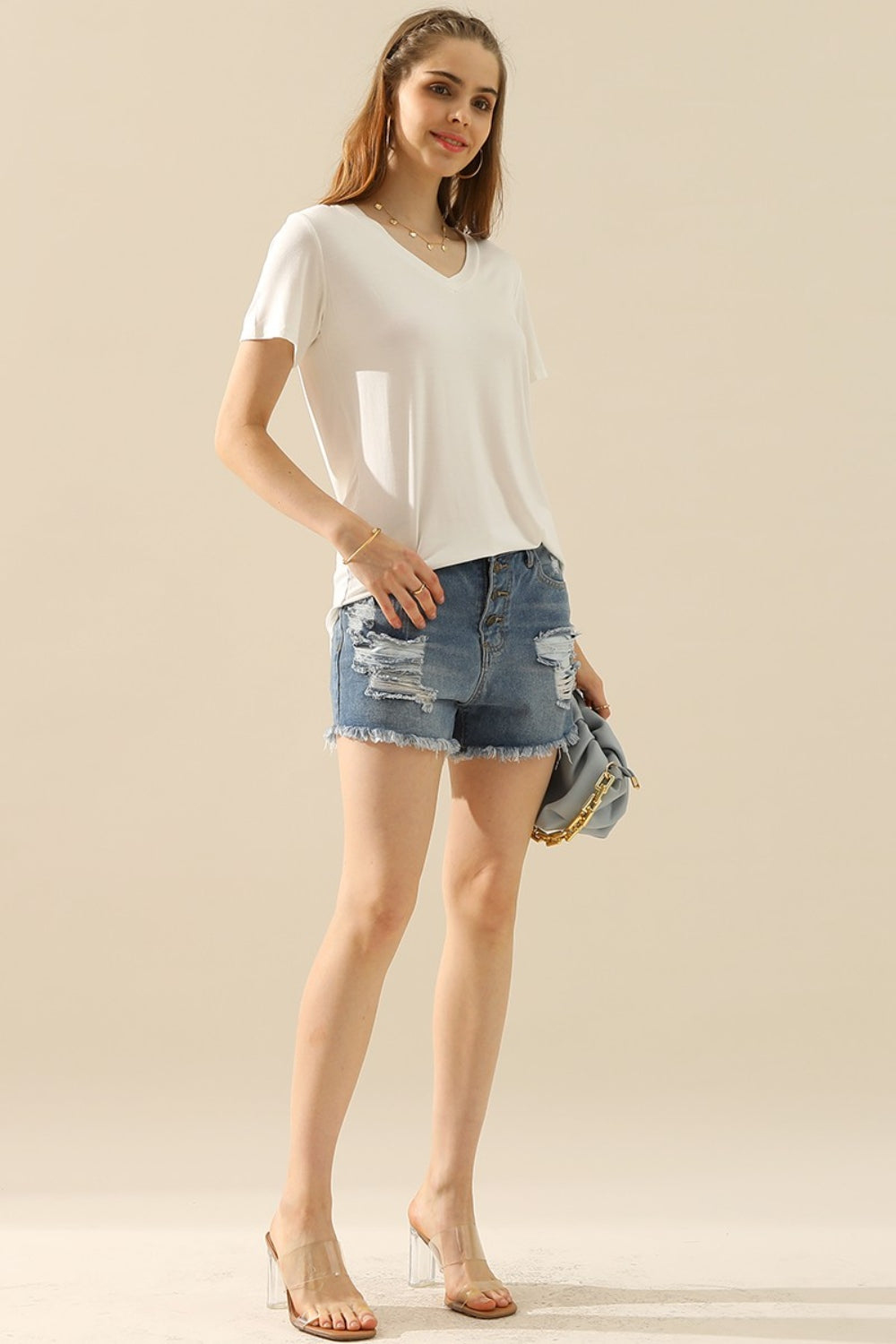 A woman with long hair wearing a Ninexis Full Size V-Neck Short Sleeve T-Shirt, a ripped denim skirt, and hoop earrings smiles at the camera.