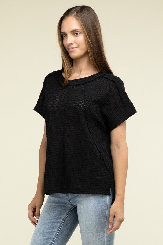 A woman with long brown hair is wearing a purple Brushed Waffle Exposed-Seam Short Sleeve Top and blue jeans, standing against a plain beige background. This casual wardrobe choice, with its short sleeves and side slits, is perfect for everyday wear.