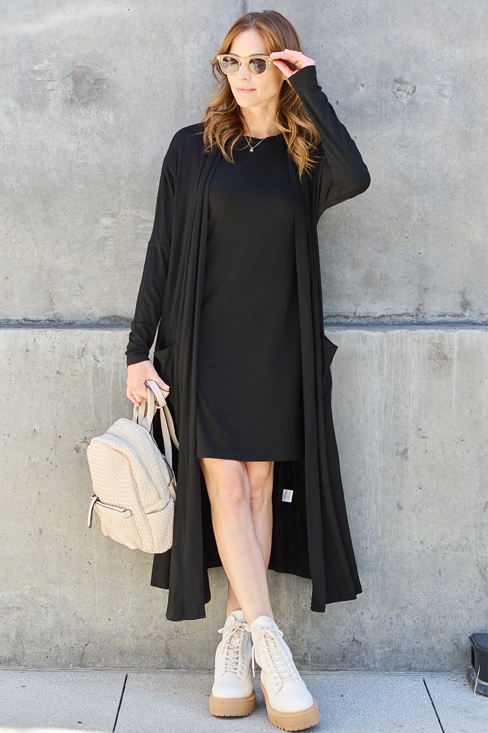 A woman stands against a concrete wall, wearing the Basic Bae Full Size Open Front Long Sleeve Cover Up made from rayon spandex, a white top, blue jeans in her perfect size, and brown shoes with her hands in her pockets. Machine wash cold for best results.