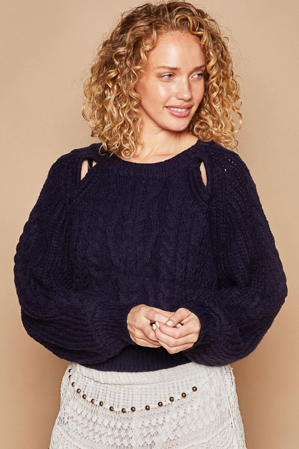 A person is wearing the POL Cable Knit Cutout Long Sleeve Sweater in navy, paired with a white crochet skirt, and is standing against a beige background.