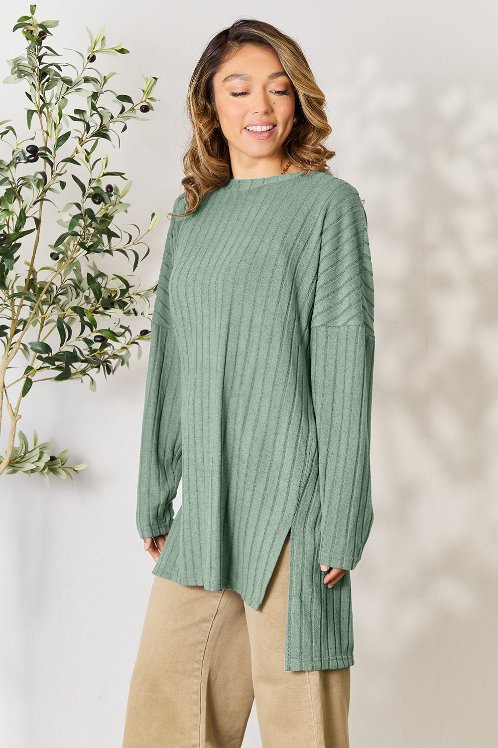 A person with shoulder-length hair smiles while wearing a beige ribbed sweater called the Basic Bae Full Size Ribbed Round Neck Long Sleeve Slit Top, along with matching beige pants, showcasing a basic style. They stand next to a green plant.