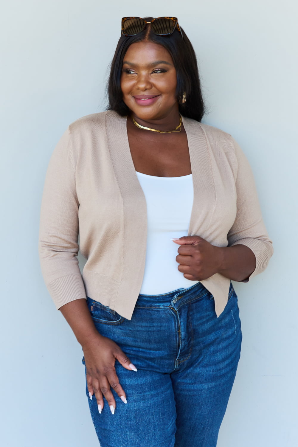 Woman with long dark hair wearing the versatile Doublju My Favorite Full Size 3/4 Sleeve Cropped Cardigan in Khaki over a white top and blue jeans, standing against a plain background.