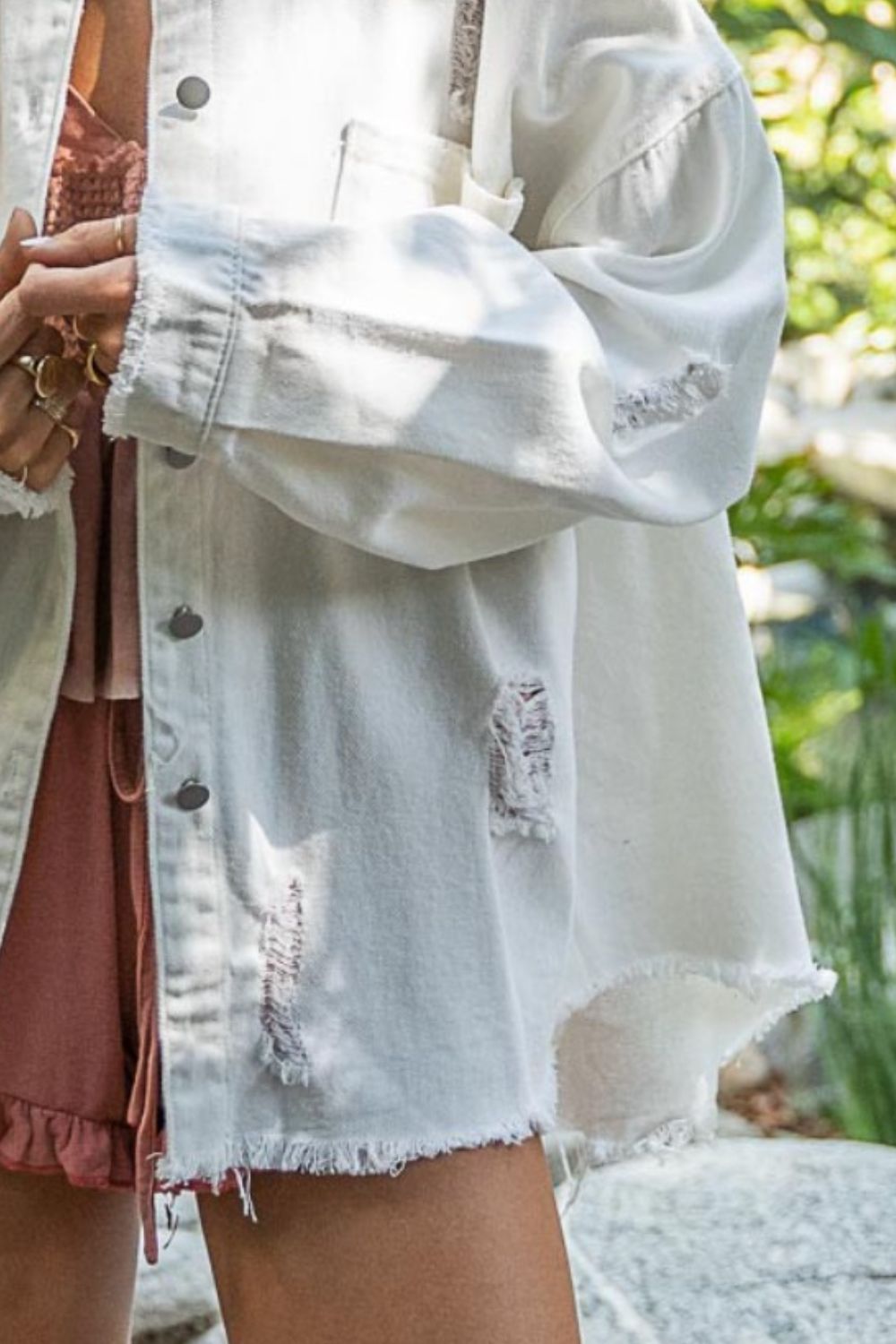 A person with long blonde hair wearing a POL Button Down Raw Hem Distressed Shacket over a peach outfit, standing outdoors in a lush garden setting.