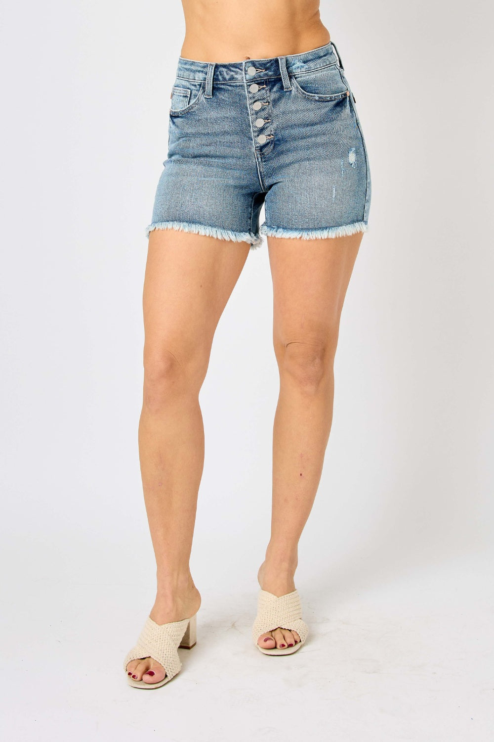 Wearing the Judy Blue Full Size Button Fly Raw Hem Denim Shorts and a white t-shirt, a person stands smiling in sandals against a plain background.