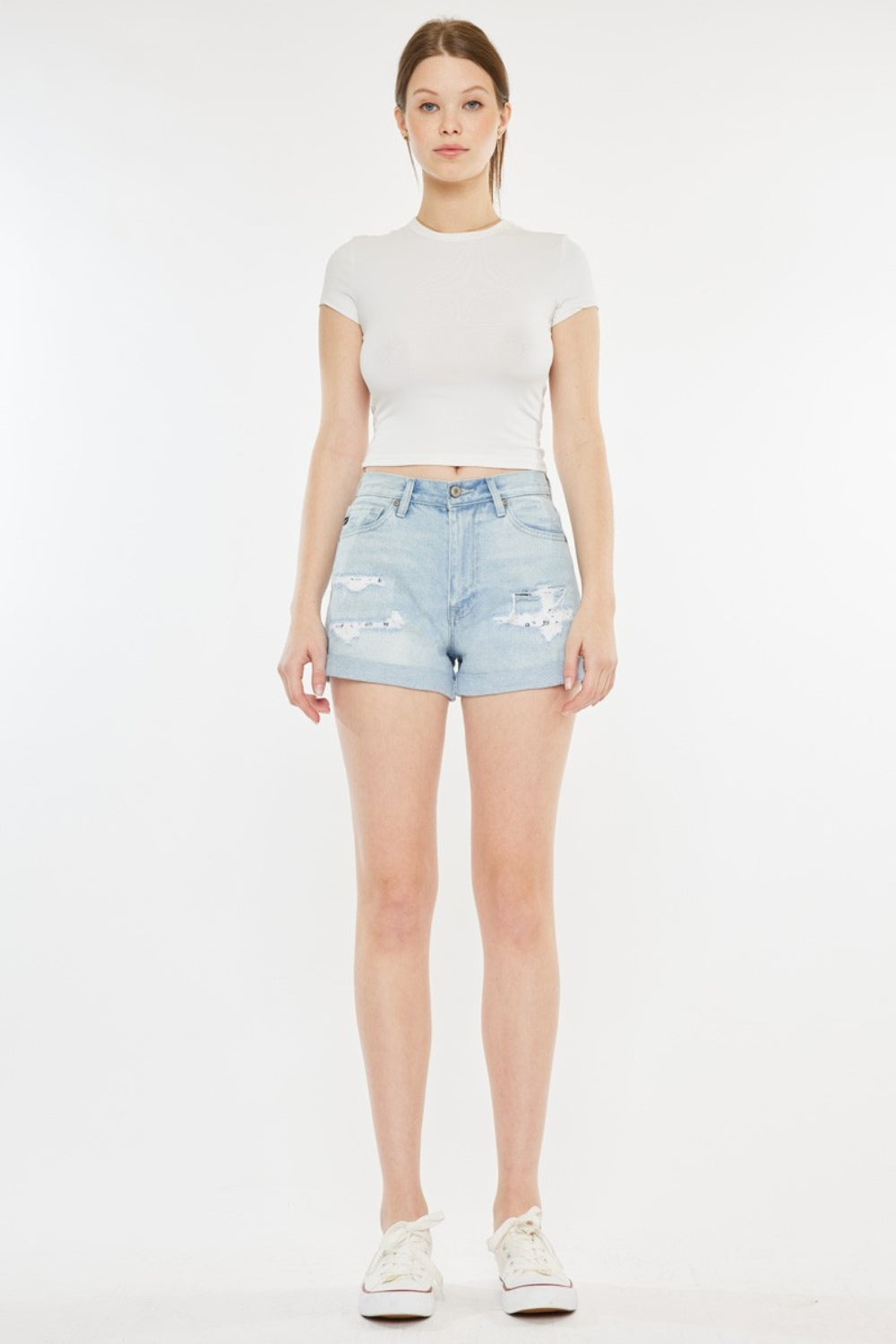 A person stands against a plain background, wearing a white short-sleeve crop top, Kancan High Rise Repaired Mom Denim Shorts in light blue, and white sneakers.